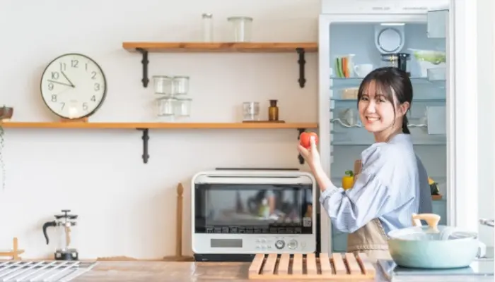 右手で冷蔵庫を開け、左手にトマトを持ちながら振り返る女性